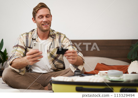 Image of smiling man packs suitcase, gets ready for holiday, buys trip from travel agency, goes on vacation, sits with credit card and laptop Image of smiling man packs suitcase, gets ready for holiday, buys trip from travel agency, goes on vacation, sits with credit card and laptop 113329494