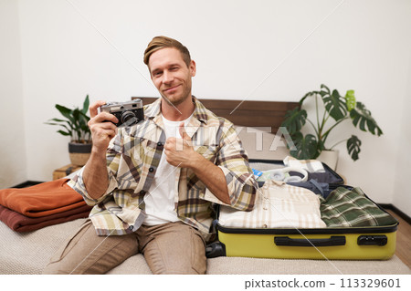 Portrait of handsome man going on vacation, packing suitcase, holding camera, travel photographer going on holiday Portrait of handsome man going on vacation, packing suitcase, holding camera, travel photographer going on holiday 113329601