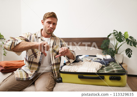 Portrait of disappointed man, tourist expresses his dislike, thumbs down, sits in hotel room with open suitcase, packing for holiday, unpacking after vacation Portrait of disappointed man, tourist expresses his dislike, thumbs down, sits in hotel room with open suitcase, packing for holiday, unpacking after vacation 113329635