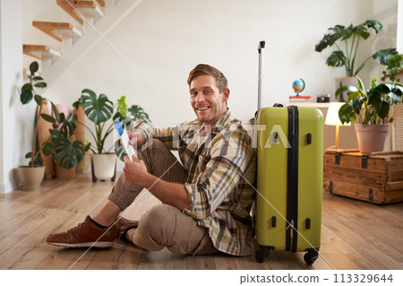 Portrait of handsome young man, tourist sitting with his suitcase, holding passport and flight tickets, going on holiday Portrait of handsome young man, tourist sitting with his suitcase, holding passport and flight tickets, going on holiday 113329644