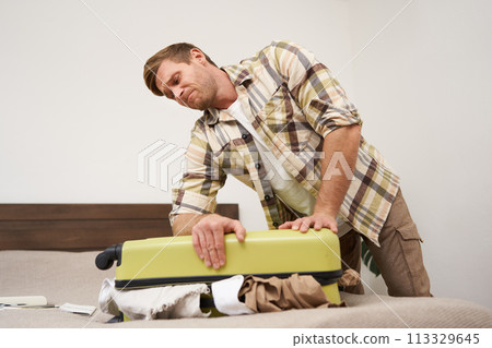 Side view of young man pushing on suitcase, trying to close it, has lots of clothes sticking out of luggage, going on long holiday, packing for vacation Side view of young man pushing on suitcase, trying to close it, has lots of clothes sticking out of luggage, going on long holiday, packing for vacation 113329645