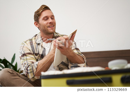 Portrait of thoughtful young man, tourist going on vacation, going through holiday check-list, writing down what he wants to take on travelling 113329669