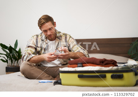Portrait of young man with suitcase, holding his plane tickets, preparing to go on holiday, packing things for vacation, tourist puts clothes in bag Portrait of young man with suitcase, holding his plane tickets, preparing to go on holiday, packing things for vacation, tourist puts clothes in bag 113329702