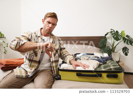 Image of young disappointed man, showing thumbs down, puts hand on his clothes inside luggage, dislike, disapprove something awful, upset with his holiday 113329703