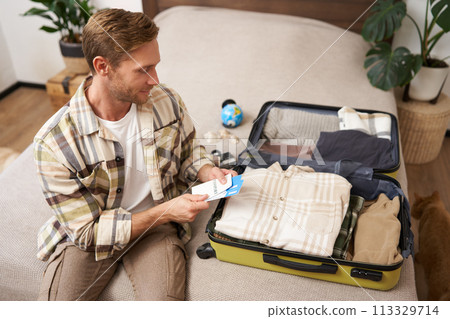 Portrait of young man going on vacation, putting his tickets in luggage, sitting with suitcase on bed, preparing for holiday or business trip Portrait of young man going on vacation, putting his tickets in luggage, sitting with suitcase on bed, preparing for holiday or business trip 113329714