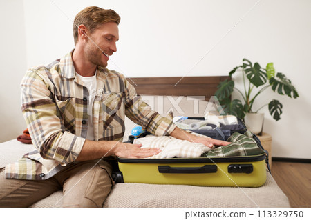 Portrait of handsome young man going on holiday, guy prepares for a trip journey, packs his suitcase, puts clothes in luggage, gets ready for adventure 113329750