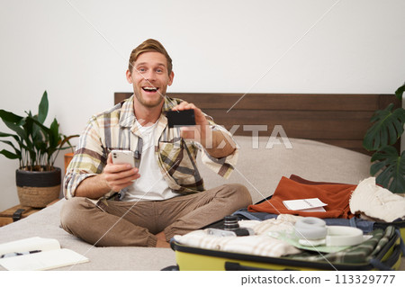 Portrait of tourist, man packing suitcase with clothes and passport, holding mobile phone, showing his credit card Portrait of tourist, man packing suitcase with clothes and passport, holding mobile phone, showing his credit card 113329777