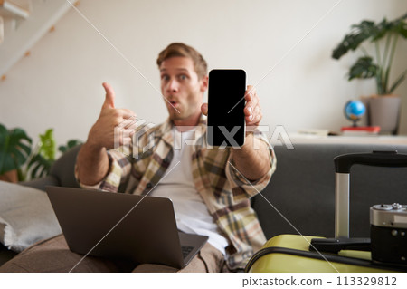 Portrait of handsome young man, showing smartphone screen, recommending mobile app for travelling or booking online, sitting with laptop and suitcase 113329812