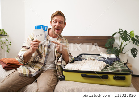 Portrait of handsome young man, traveller shows his passport and two tickets, packing suitcase with clothes, going on vacation Portrait of handsome young man, traveller shows his passport and two tickets, packing suitcase with clothes, going on vacation 113329820