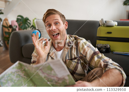 Smiling handsome tourist, man with road travel map, sits on floor near suitcase, shows okay, ok sign, recommending a tour Smiling handsome tourist, man with road travel map, sits on floor near suitcase, shows okay, ok sign, recommending a tour 113329938