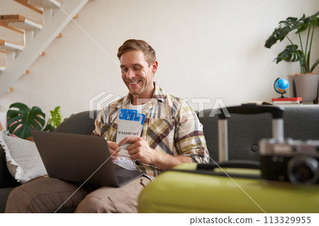 Portrait of happy tourist, man looking excited at laptop screen, holding flight tickets, going on vacation, excited about holiday, sits with suitcase in living room 113329955