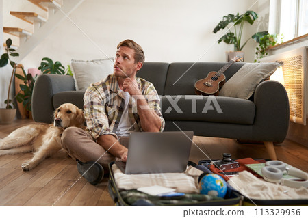Portrait of man with dog, sitting on floor, has suitcase and laptop opened, booking or buying online, planning holiday, packing for vacation 113329956