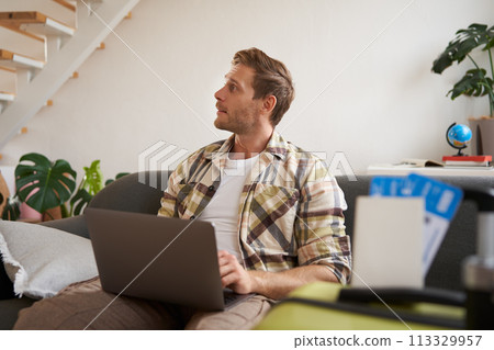 Portrait of young handsome man sitting with laptop, has passport and two plane tickets prepared, planning his journey on computer, booking hotels and restaurants online for the trip 113329957