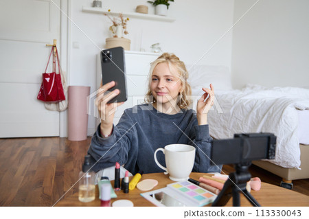 Portrait of young beauty blogger, recording video on digital camera, doing online live stream while applying makeup, talking to her followers, sitting on bedroom floor Portrait of young beauty blogger, recording video on digital camera, doing online live stream while applying makeup, talking to her followers, sitting on bedroom floor 113330043