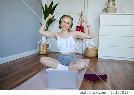 Portrait of fit and healthy, sporty woman showing her muscles on camera, wearing wireless headphones, using laptop for home workout training Portrait of fit and healthy, sporty woman showing her muscles on camera, wearing wireless headphones, using laptop for home workout training 113330124