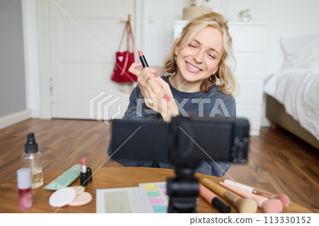 Portrait of young creative social media content creator, woman showing lipstick swatches on her hand, recording video about beauty and makeup, sitting in her room in front of digital camera 113330152