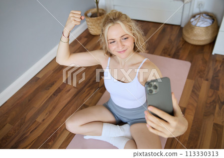 Portrait of young sporty woman taking selfie during workout training, content creator takes photos, shows muscles on camera, flexing biceps 113330313