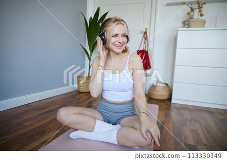 Young and sporty woman working out at home, doing fitness training alone on yoga mat, listens to music in wireless headphones Young and sporty woman working out at home, doing fitness training alone on yoga mat, listens to music in wireless headphones 113330349