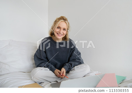 Portrait of young beautiful woman sitting on bed with laptop and notebooks, working from home, freelancing. Female student studying online, video chats, connects to lesson remotely Portrait of young beautiful woman sitting on bed with laptop and notebooks, working from home, freelancing. Female student studying online, video chats, connects to lesson remotely 113330355