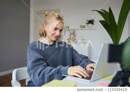 Portrait of young blond woman, female college student works from home on assignment, uses laptop, studies remotely, sits in room in front of computer Portrait of young blond woman, female college student works from home on assignment, uses laptop, studies remotely, sits in room in front of computer 113330419