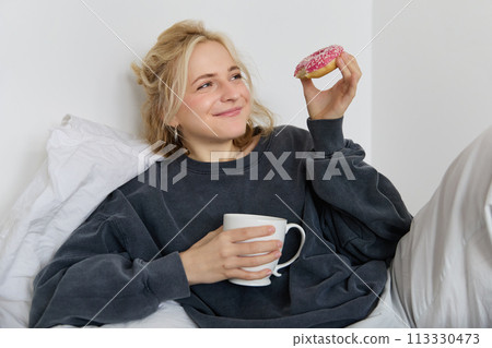Close up portrait of beautiful smiling, blond woman, lying in bed, drinks tea and eats doughnut on a lazy weekend or day-off Close up portrait of beautiful smiling, blond woman, lying in bed, drinks tea and eats doughnut on a lazy weekend or day-off 113330473