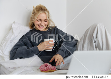 Portrait of happy blond girl, female model lying in bed in cosy clothes, eating and watching movie on laptop, smiling while looking at screen Portrait of happy blond girl, female model lying in bed in cosy clothes, eating and watching movie on laptop, smiling while looking at screen 113330613
