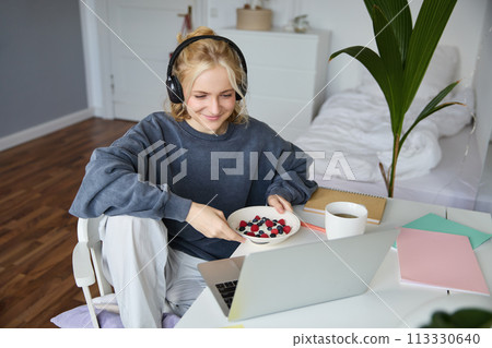 Portrait of smiling young woman, watching tv show in headphones, eating breakfast and looking at laptop screen Portrait of smiling young woman, watching tv show in headphones, eating breakfast and looking at laptop screen 113330640