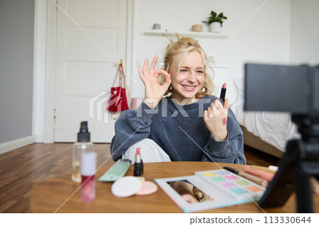 Cheerful, smiling young woman recording a vlog, showing okay, ok gesture and favorite lipstick, recommending beauty product for social media followers, using digital camera to create content 113330644