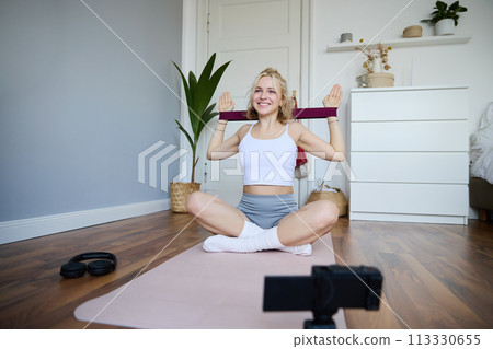 Portrait of young blond woman, social media vlogger using digital camera during workout, shooting video about exercises and fitness, stretching with rubber resistance band 113330655
