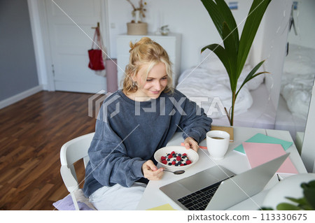 Portrait of smiling blond young woman, eating in front of laptop, watching videos online while having breakfast, enjoying dessert, sitting in bedroom 113330675