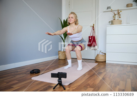 Portrait of young woman, fitness instructor making a video for sport vlog, doing squats on camera, using resistance band, working out at home 113330689