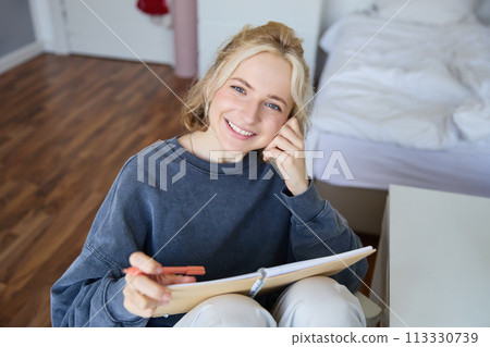 Portrait of smiling, beautiful young candid woman, sitting in bedroom with pen and notebook, writing in journal, making notes, looking happy at camera Portrait of smiling, beautiful young candid woman, sitting in bedroom with pen and notebook, writing in journal, making notes, looking happy at camera 113330739