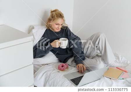 Portrait of young woman, student studying in her bed, relaxing while preparing homework, eating doughnut, using laptop in bedroom and drinking tea 113330775