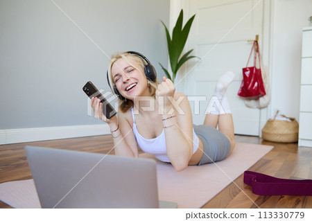 Young woman doing fitness workout at home, lying on rubber yoga mat, holding smartphone, wearing headphones, using laptop to watch sports videos Young woman doing fitness workout at home, lying on rubber yoga mat, holding smartphone, wearing headphones, using laptop to watch sports videos 113330779
