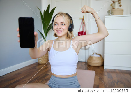 Portrait of beautiful and fit young woman, staying healthy working out at home, showing mobile app, blank screen, flexing biceps, recommending smartphone application for exercises 113330785