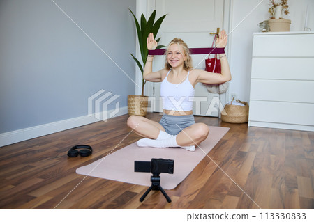 Portrait of young smiling woman, fitness instructor recording video about workout, showing how to exercise at home and use rubber resistance band, sitting on yoga mat 113330833