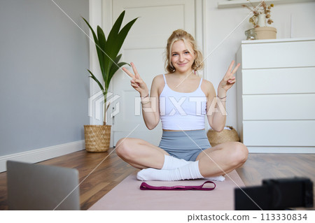 Portrait of cute blond woman shows peace sign, smiles, records yoga vlog on digital camera, workout at home on rubber mat in room 113330834