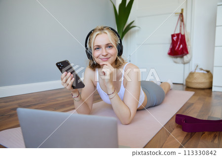 Portrait of young sporty woman, connects to online workout training session, doing exercises, lying on rubber mat, listens to fitness instructor in headphones, holding mobile phone 113330842