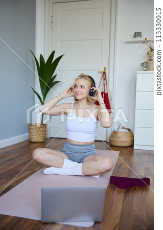 Vertical shot of smiling young woman using video tutorials for workout at home, sitting with laptop in wireless headphones on yoga mat, follows fitness instructor on social media, repeats exercises Vertical shot of smiling young woman using video tutorials for workout at home, sitting with laptop in wireless headphones on yoga mat, follows fitness instructor on social media, repeats exercises 113330915