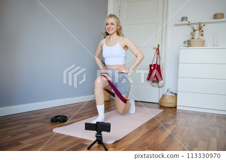 Portrait of fitness instructor, young sporty vlogger woman doing exercises on camera, shooting video about workout, using resistance band and doing sit-ups at home in empty room Portrait of fitness instructor, young sporty vlogger woman doing exercises on camera, shooting video about workout, using resistance band and doing sit-ups at home in empty room 113330970
