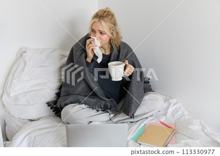 Portrait of young woman sitting at home sick, feeling unwell, drinking tea, spending time in bed, has runny nose, working on laptop Portrait of young woman sitting at home sick, feeling unwell, drinking tea, spending time in bed, has runny nose, working on laptop 113330977
