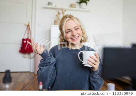 Portrait of cute smiling woman, blond girl drinks tea and records a casual, lifestyle video blog, vlogger sits in a room with camera and stabiliser, holds cup of coffee and talks Portrait of cute smiling woman, blond girl drinks tea and records a casual, lifestyle video blog, vlogger sits in a room with camera and stabiliser, holds cup of coffee and talks 113330979