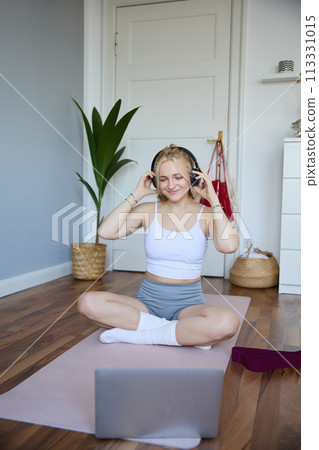 Portrait of young woman at home, connects to online gym training session, following fitness instructions on laptop, wearing wireless headphones during workout 113331015