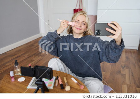 Portrait of beautiful young lifestyle blogger, woman vlogger recording video on digital camera, live stream makeup tutorial on smartphone app, sitting in bedroom, using brush on her cheeks 113331016