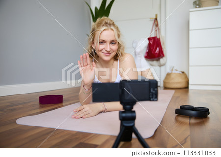 Portrait of smiling blond woman saying hello to her followers, fitness instructor recording video of herself showing workout exercises at home, lying on rubber yoga mat, looking at digital camera 113331033