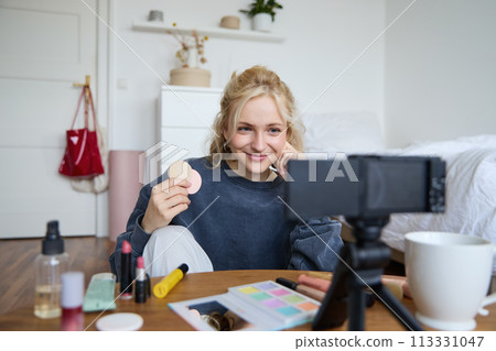 Image of young woman, vlogger records video on digital camera, shows beauty products, recommending makeup for audience on social media, sits on front in a room Image of young woman, vlogger records video on digital camera, shows beauty products, recommending makeup for audience on social media, sits on front in a room 113331047