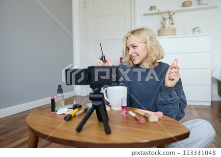 Portrait of young smiling woman in her room, recording video on camera, lifestyle vlog for social media, holding mascara, reviewing her makeup beauty products, showing how to use cosmetics Portrait of young smiling woman in her room, recording video on camera, lifestyle vlog for social media, holding mascara, reviewing her makeup beauty products, showing how to use cosmetics 113331048