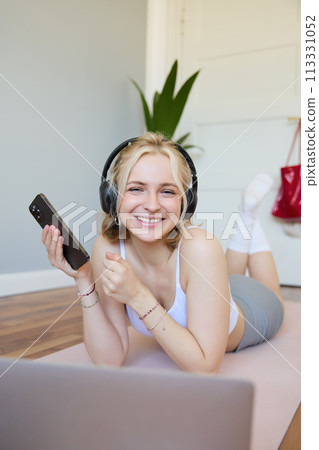Vertical shot of young blond woman in headphones, lying on rubber mat, using laptop, holding mobile phone, listens to music or workout podcast, doing exercises at home 113331052