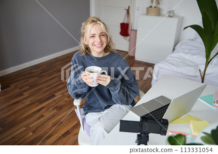 Image of young woman, social media influencer, editing her video on laptop, sits in a room with computer and digital camera, drinking coffee, smiling at camera 113331054