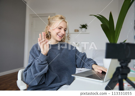 Portrait of young social media content creator, woman sits in room with laptop and digital camera, records lifestyle vlog, says hello, chats to someone online via computer 113331055